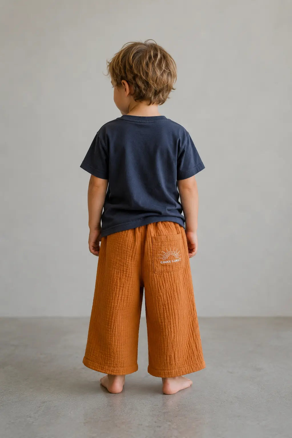 Back view of boy wearing Cooee Coast Ochre coastal dune pants featuring embroidered back pocket, relaxed fit.